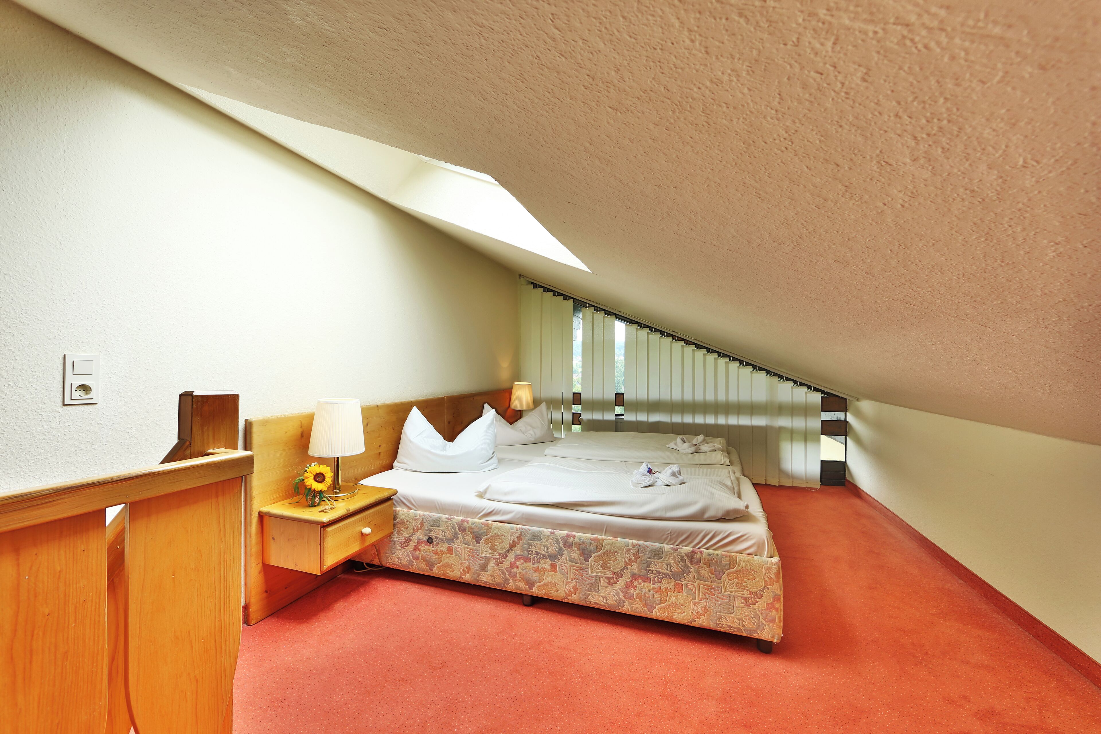 Un dormitorio acogedor en el ático, con una cama individual, una mesita de noche, una lámpara y una ventana con persianas.