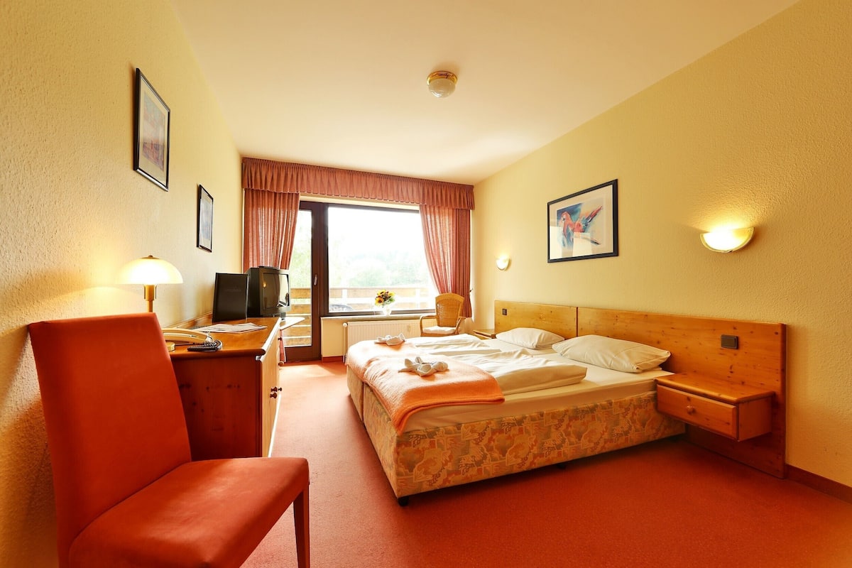 A hotel room with a bed, a desk with a chair, a lamp, a window with curtains, and a painting on the wall.