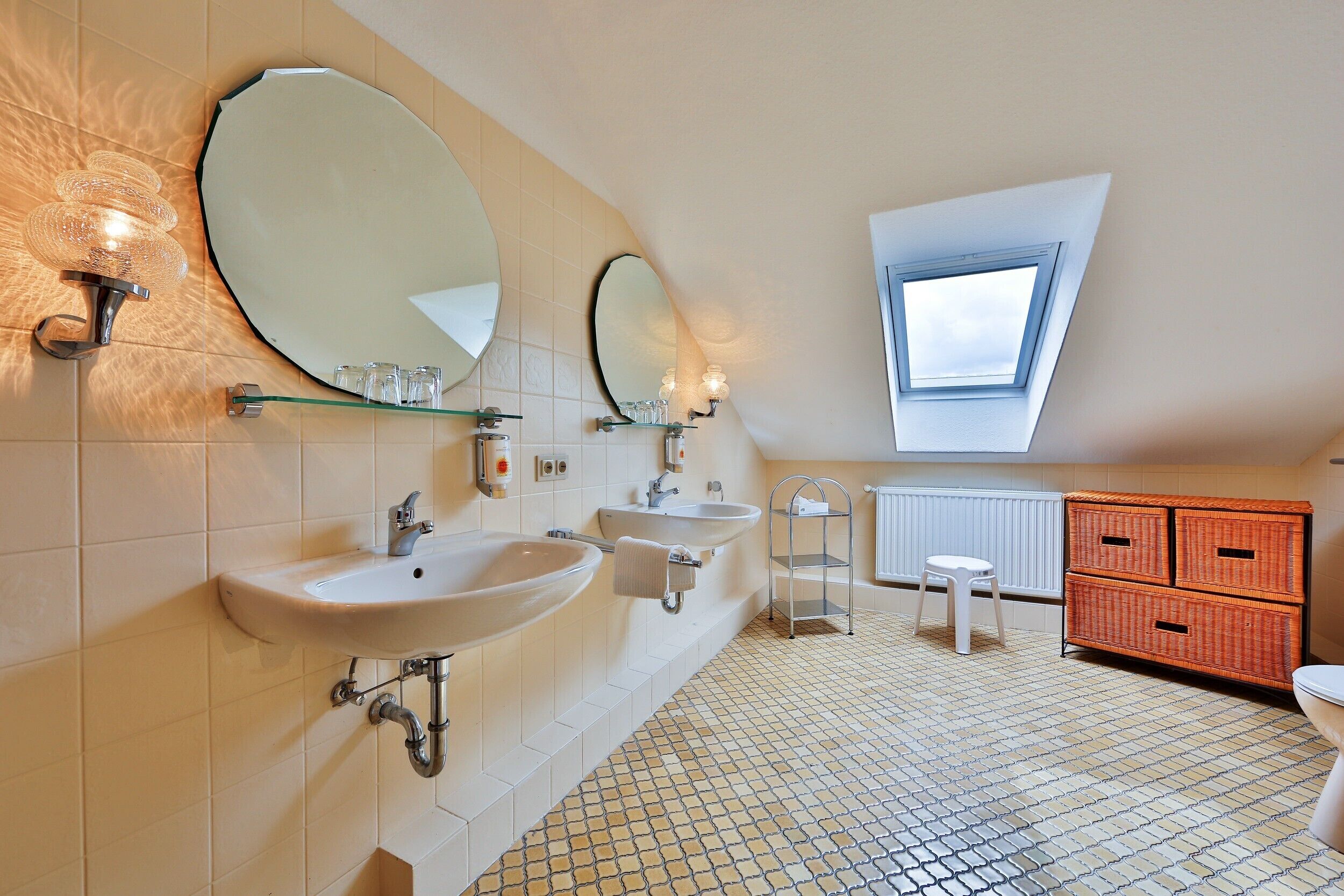 Une salle de bain avec deux lavabos, un grand miroir, une étagère en verre, un tabouret, une fenêtre et un sol carrelé.