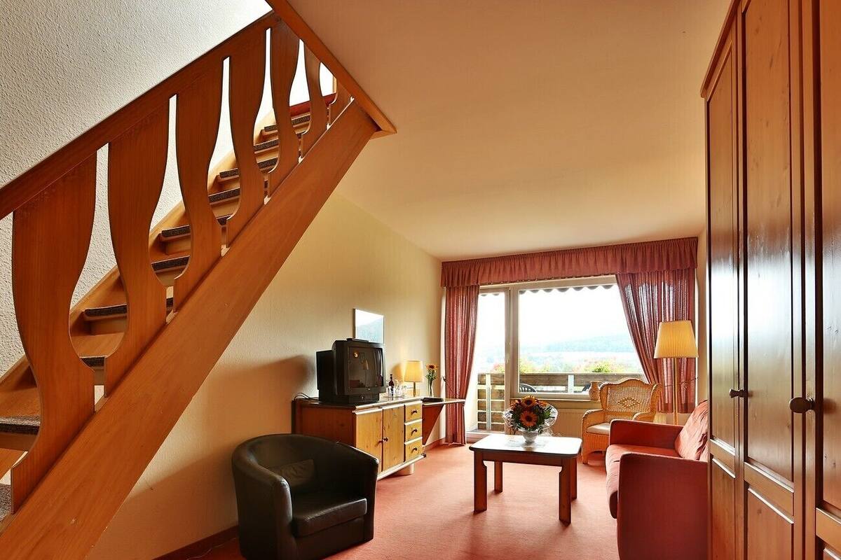 A hotel room with a wooden staircase, a television on a wooden cabinet, a sofa, a table with a vase, and a view of the outdoors.