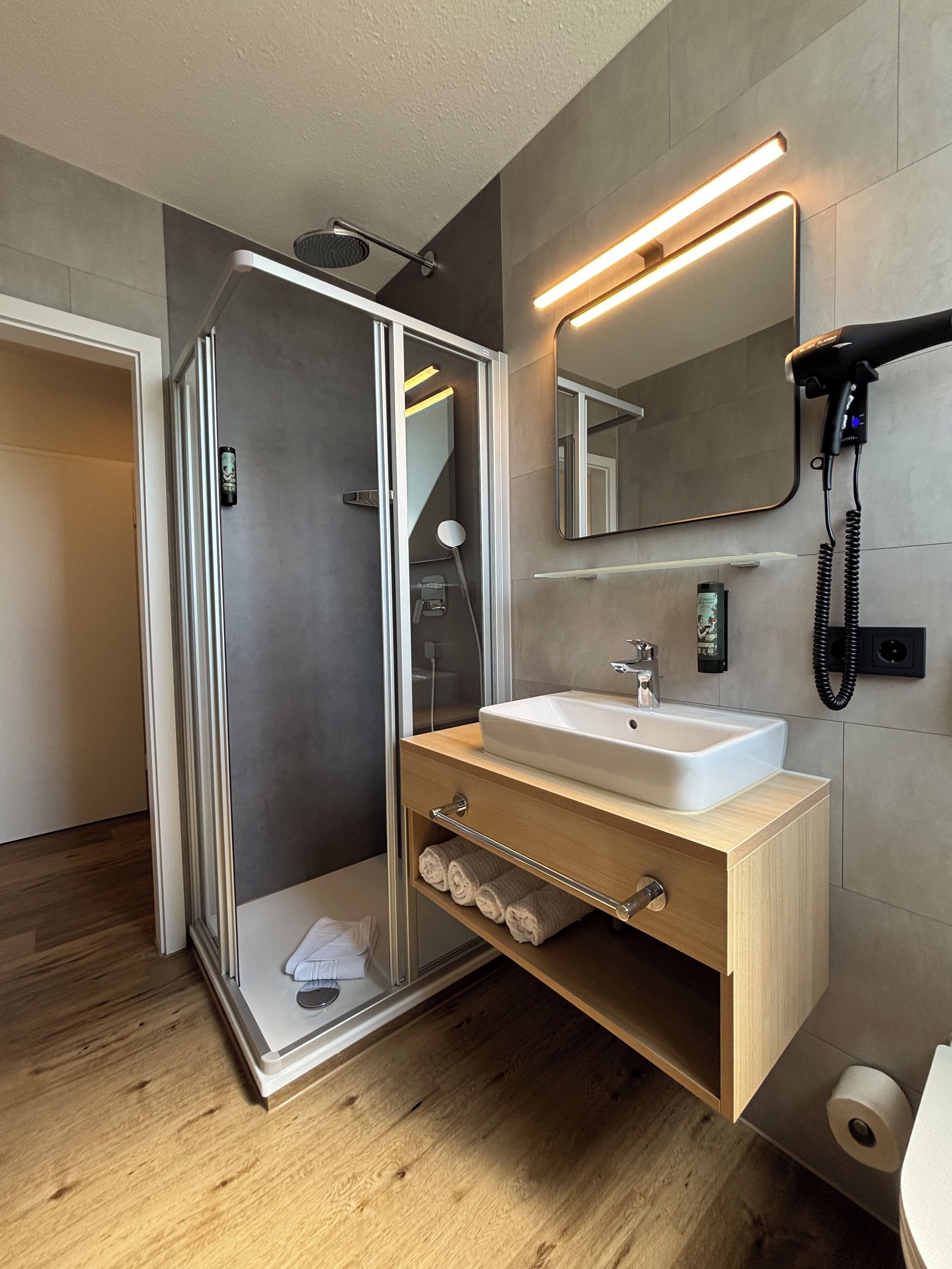 Un baño moderno con ducha envuelta en vidrio, tocador de madera con lavamanos y un espejo sobre el tocador.