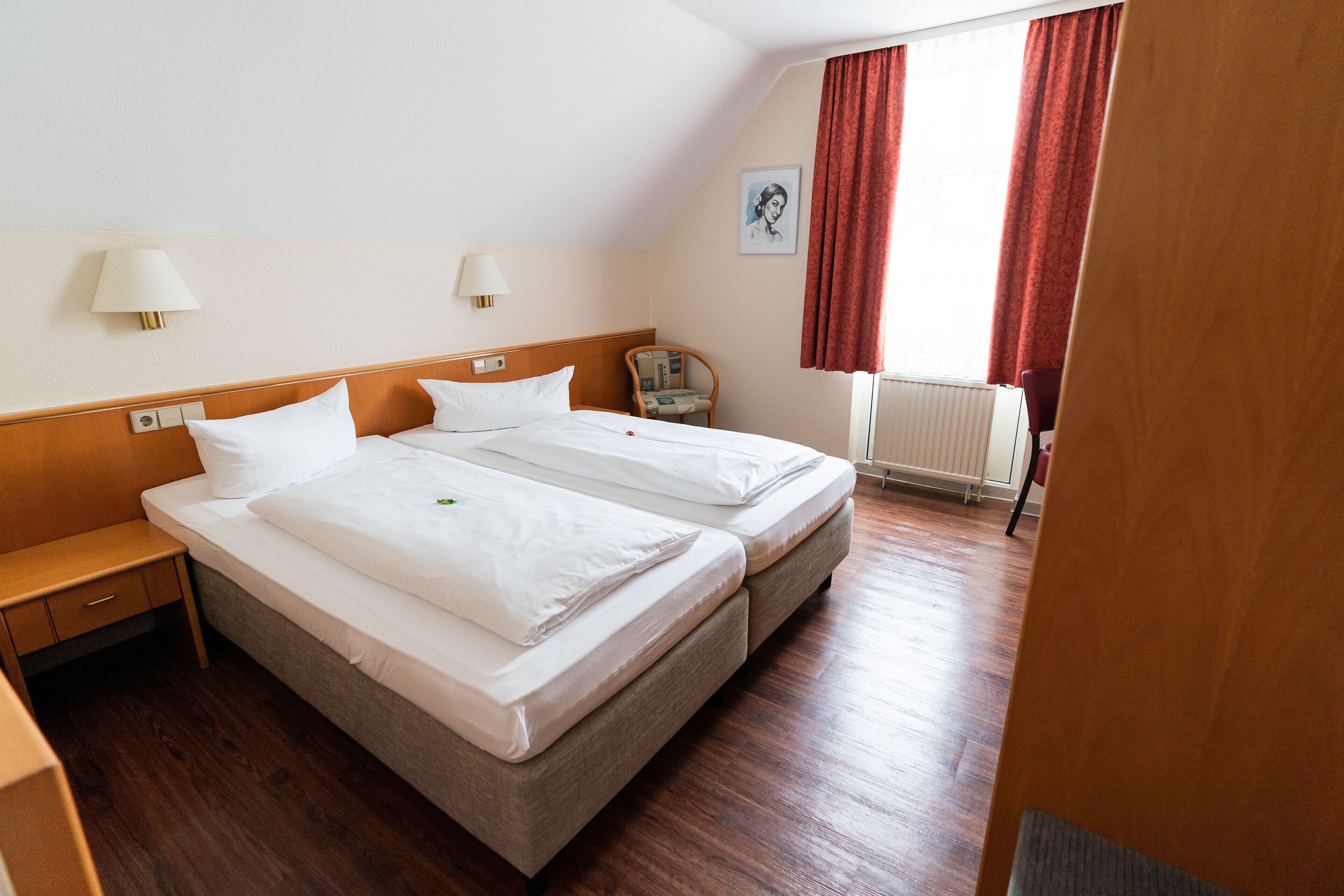 A hotel room with two beds, wooden headboards, a nightstand, a chair, and a window with red curtains.