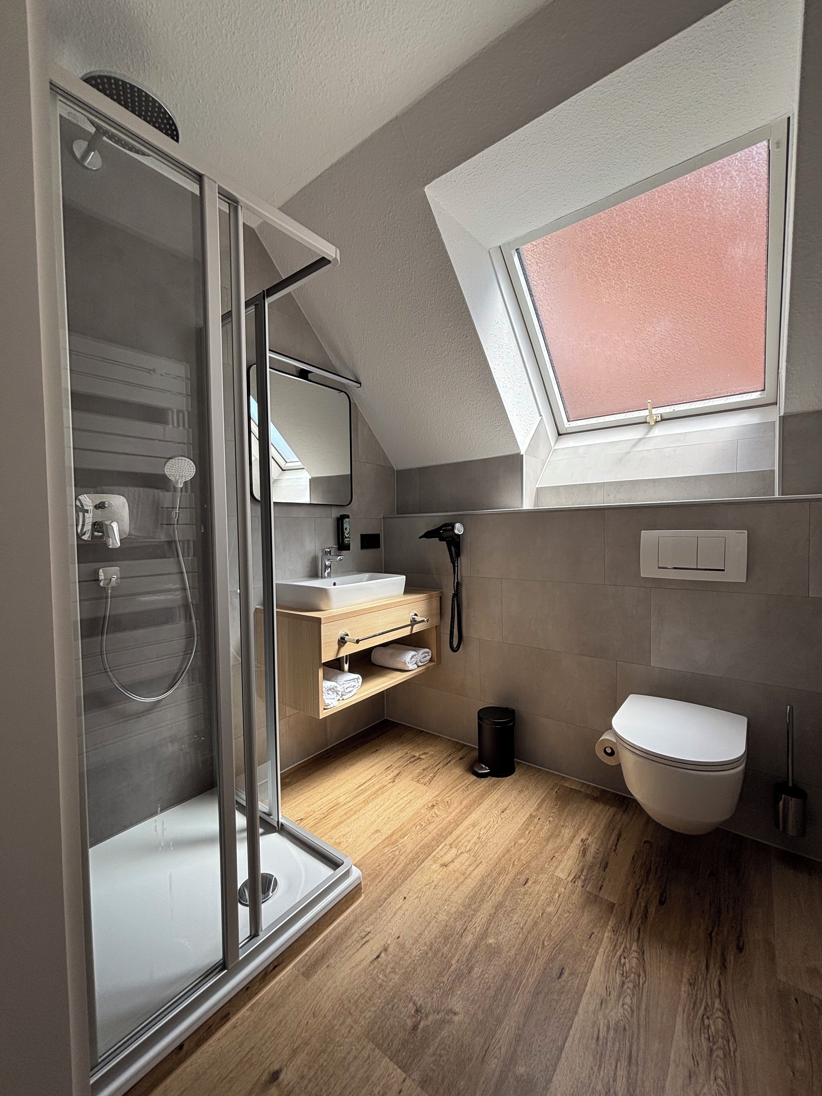 A modern bathroom with a glass shower enclosure, a wooden vanity, a white toilet, and a skylight.