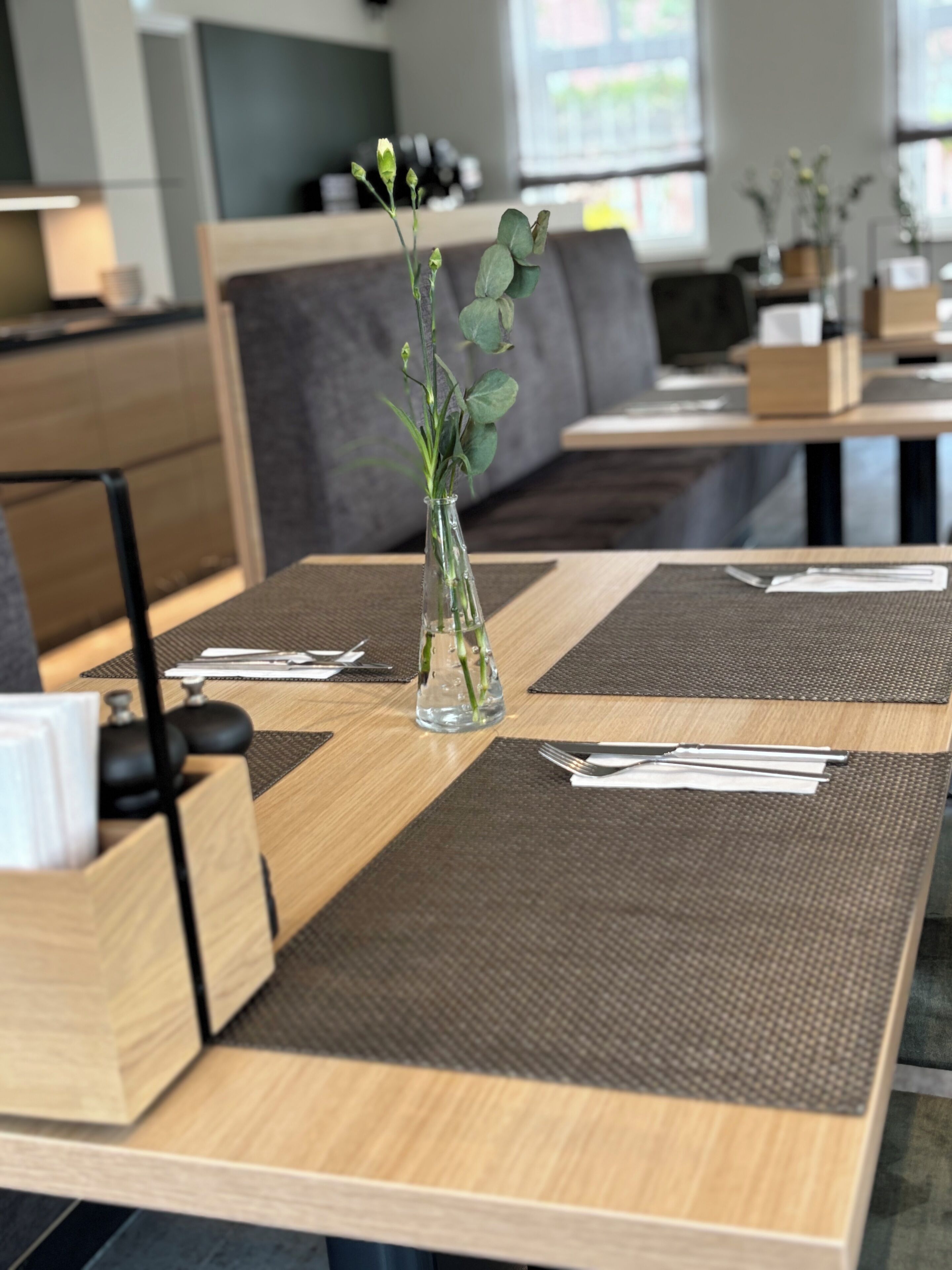 Una mesa de restaurante con un florero de vidrio transparente que contiene plantas verdes, cubiertos y una carta.