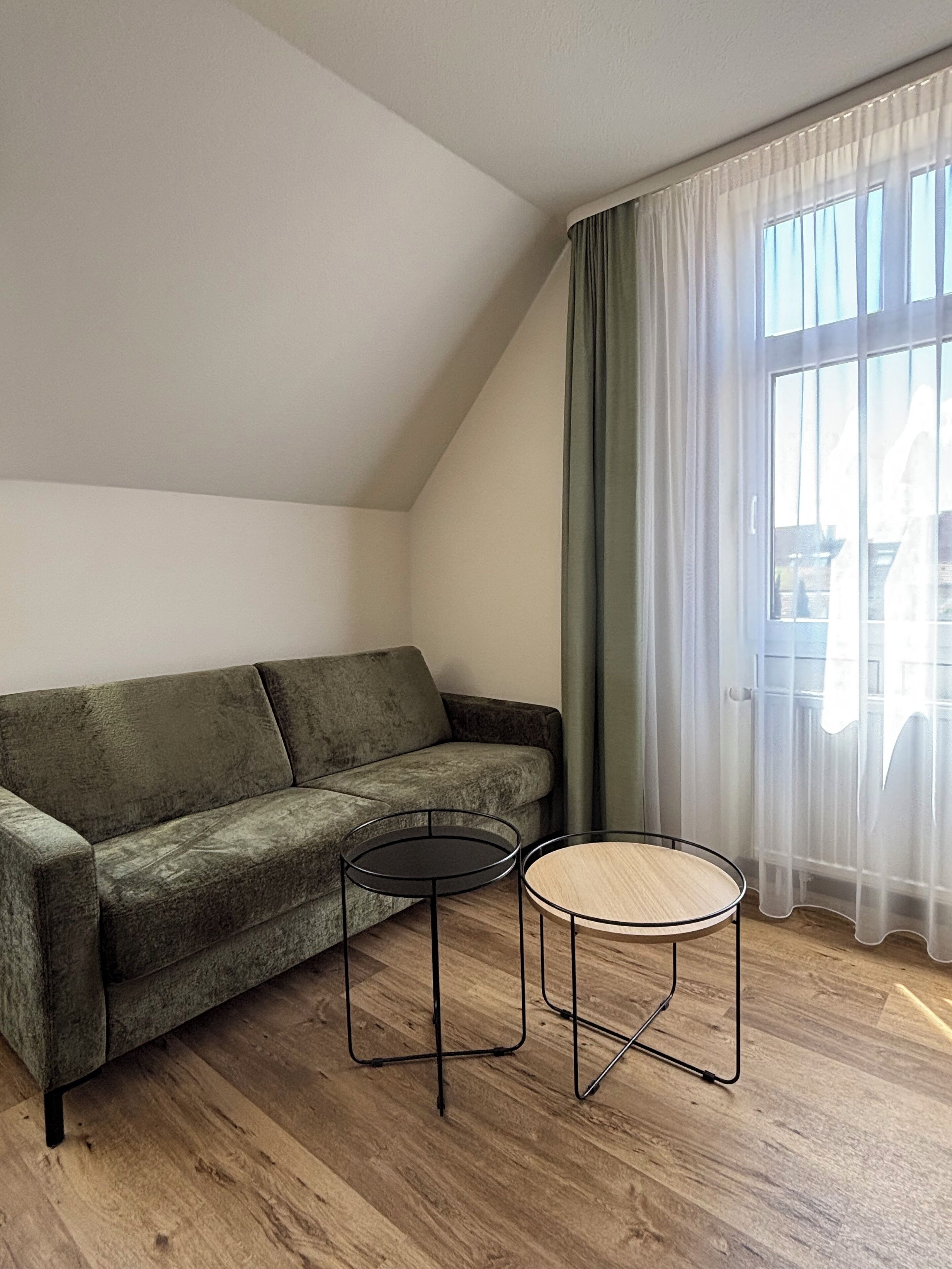A modern living room with a green sofa, two small round tables, and a large window with sheer curtains.