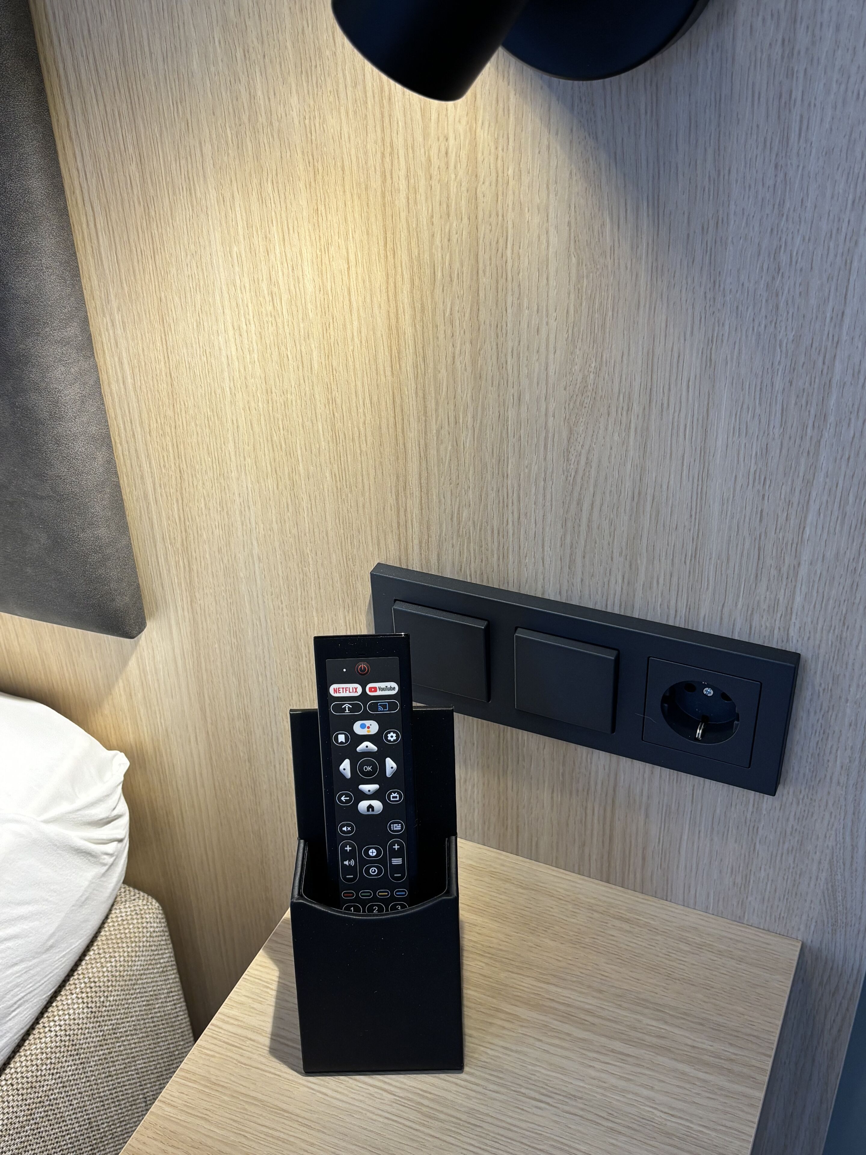 A black remote control in a holder on a wooden bedside table next to a bed.
