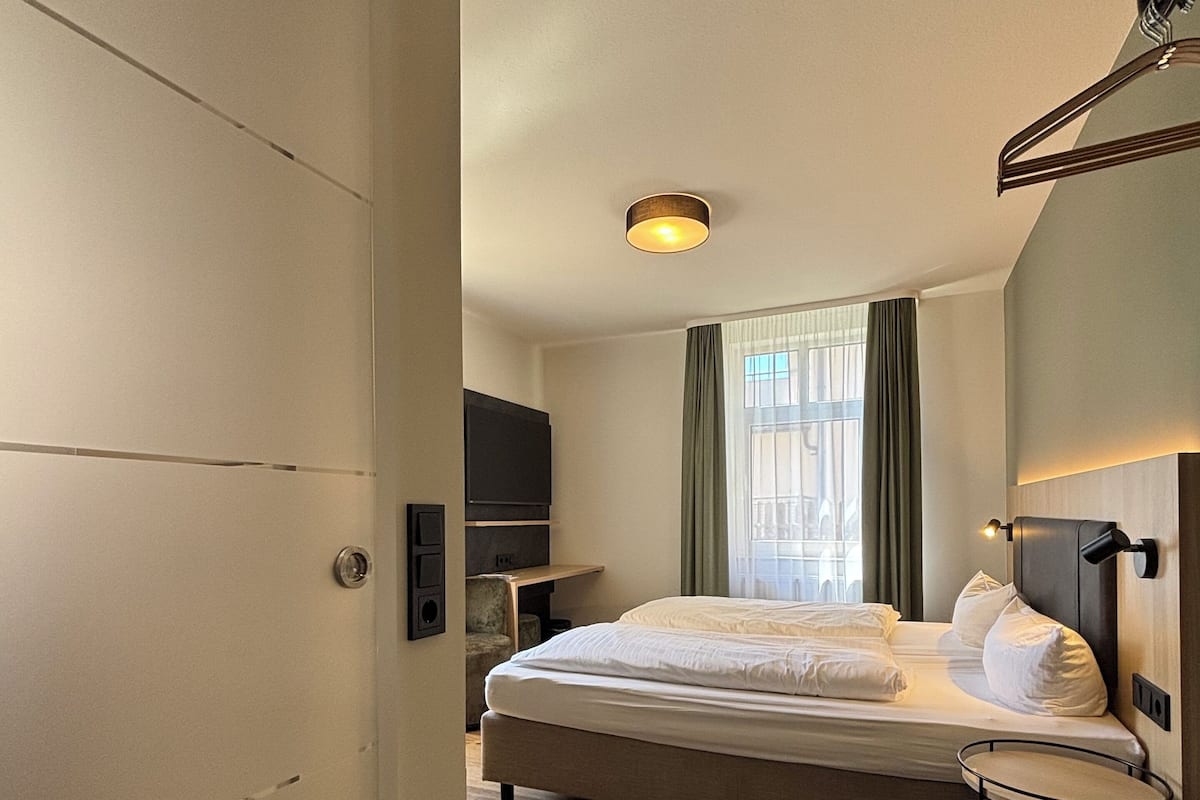 A hotel room with a bed, a TV, a bedside table, and a window with curtains.