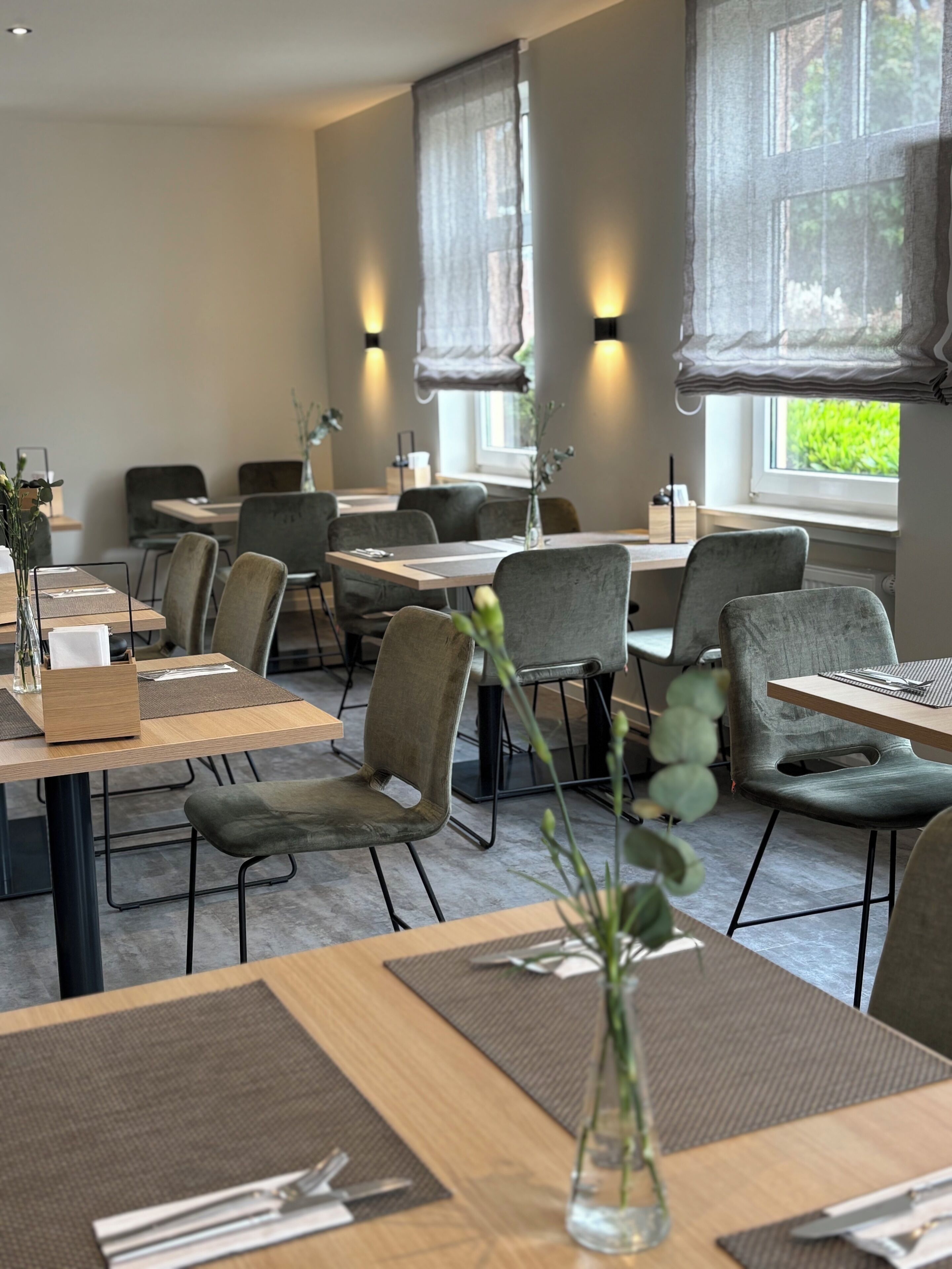 A modern dining area with wooden tables and grey upholstered chairs, large windows with sheer curtains, and a view of greenery outside.