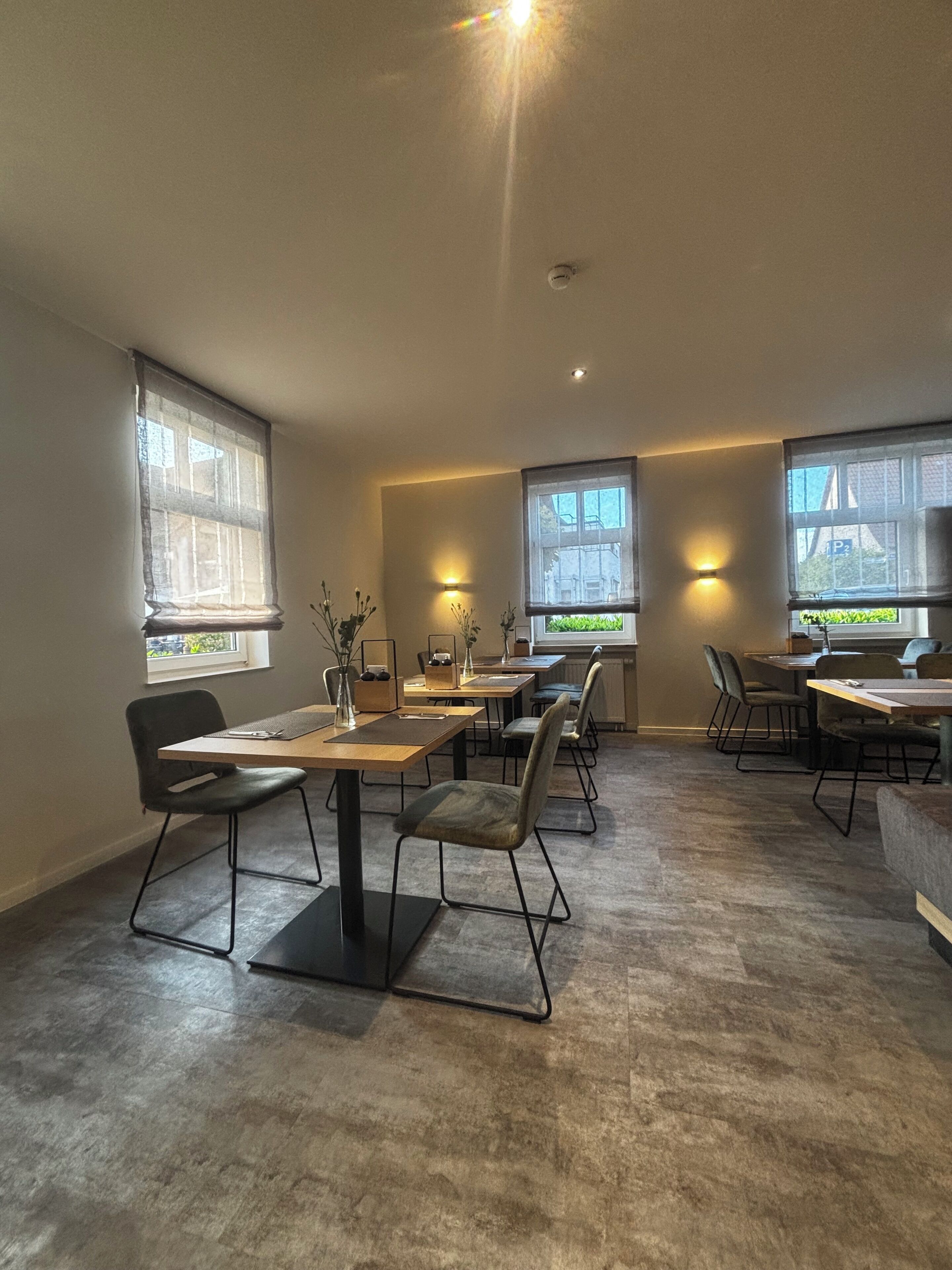 A modern dining area with multiple tables and chairs, large windows with views, and recessed ceiling lighting.