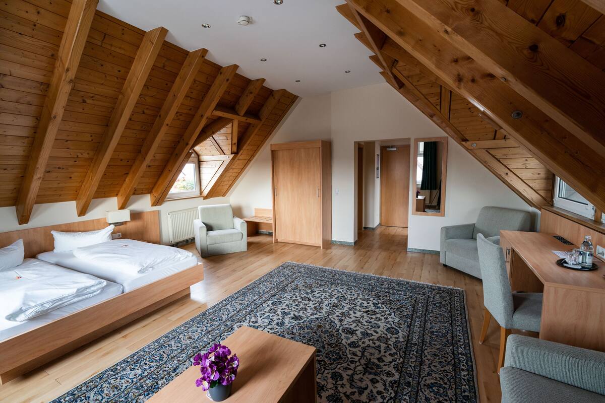 A spacious loft bedroom with a large bed, a desk, and a chair. There is a wooden ceiling with exposed beams and a window providing natural light.