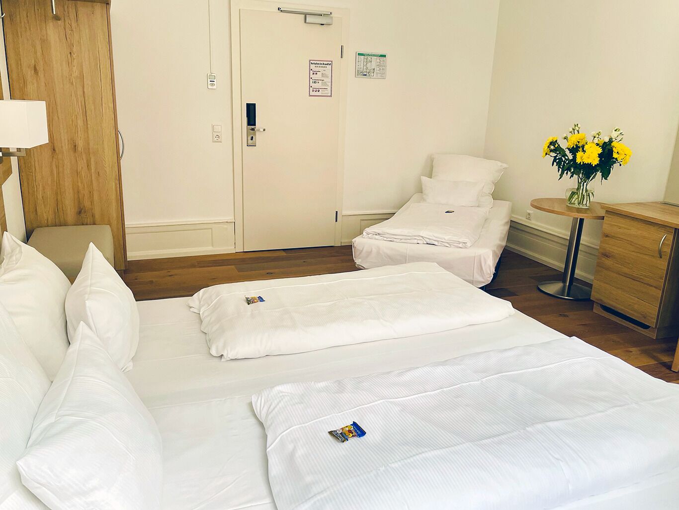 A hotel room with two beds, a wooden wardrobe, a small table with a vase of flowers, and a wall-mounted clock.