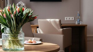 A hotel room with a round dining table, a vase of flowers, a flat-screen TV mounted on the wall, a chair, and a small side table with a bottle.
