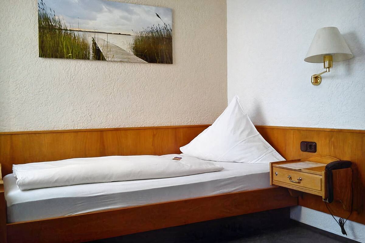 Un lit simple avec une tête de lit en bois, une table de chevet avec une lampe et un tableau représentant un paysage accroché au mur.