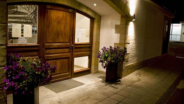 A hotel entrance with a wooden double door, a stone wall, and two potted plants with purple flowers.