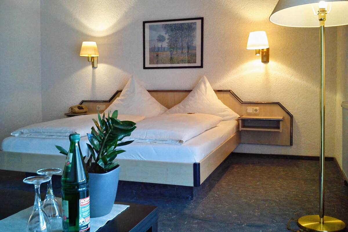 Une chambre d’hôtel avec un lit, des lampes de chevet, une table de chevet, une plante en pot et une table avec des verres et une bouteille.
