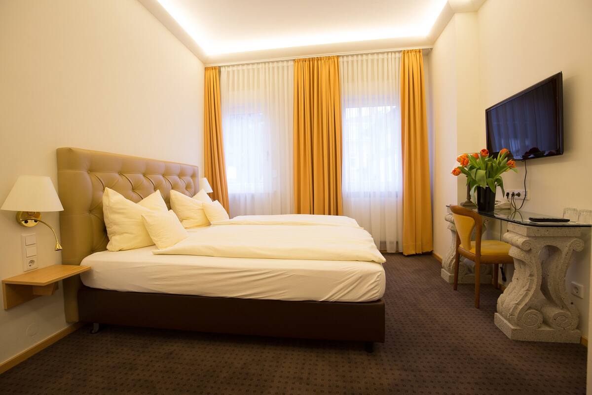 A hotel room with a large bed, a desk with a chair, a flat-screen TV, and a vase with flowers.