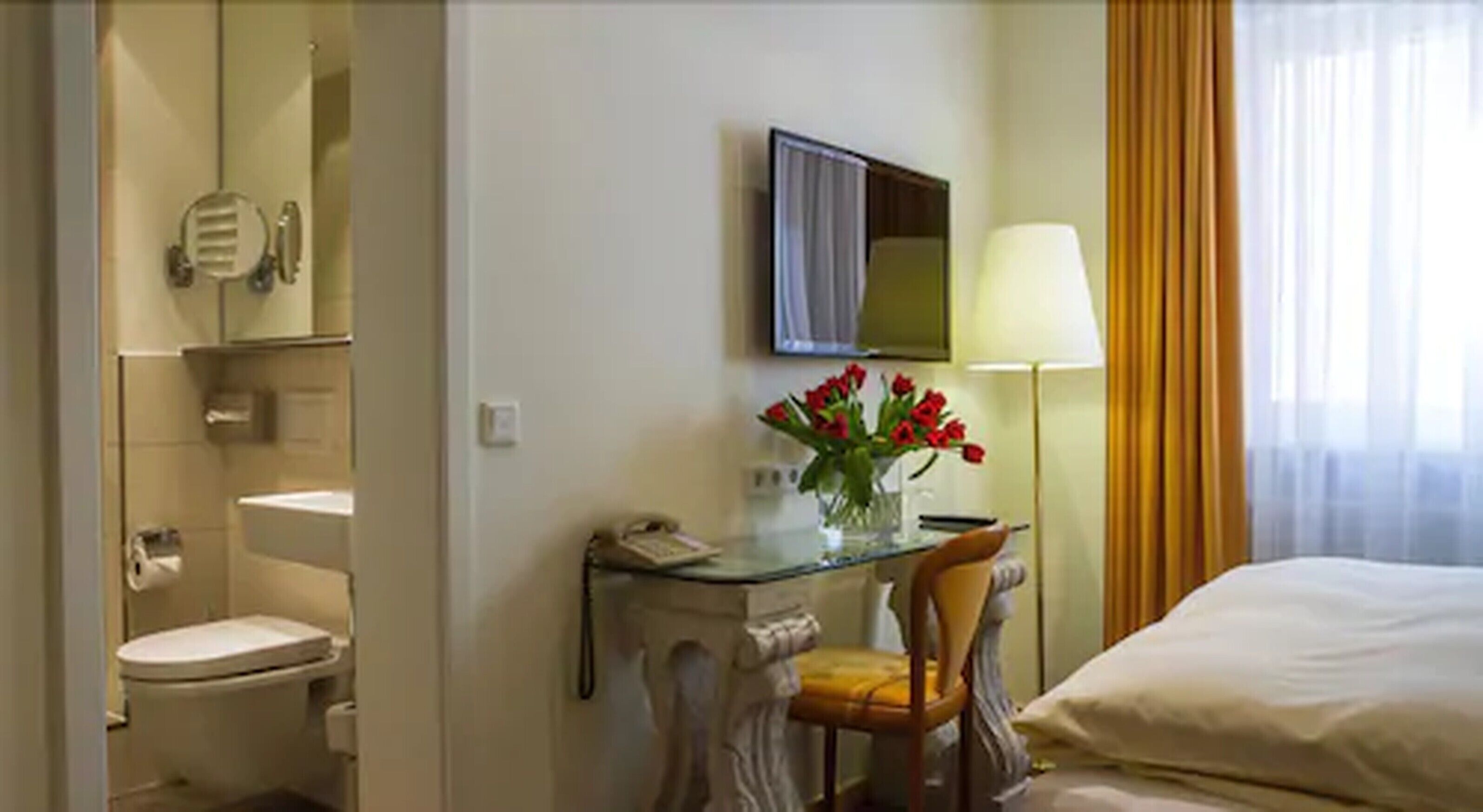A hotel room with a bed, a desk with a lamp, a chair, a toilet, and a mirror.