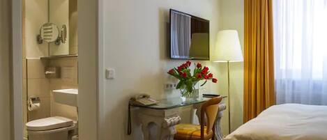 A hotel room with a bed, a desk with a lamp, a chair, a toilet, and a mirror.