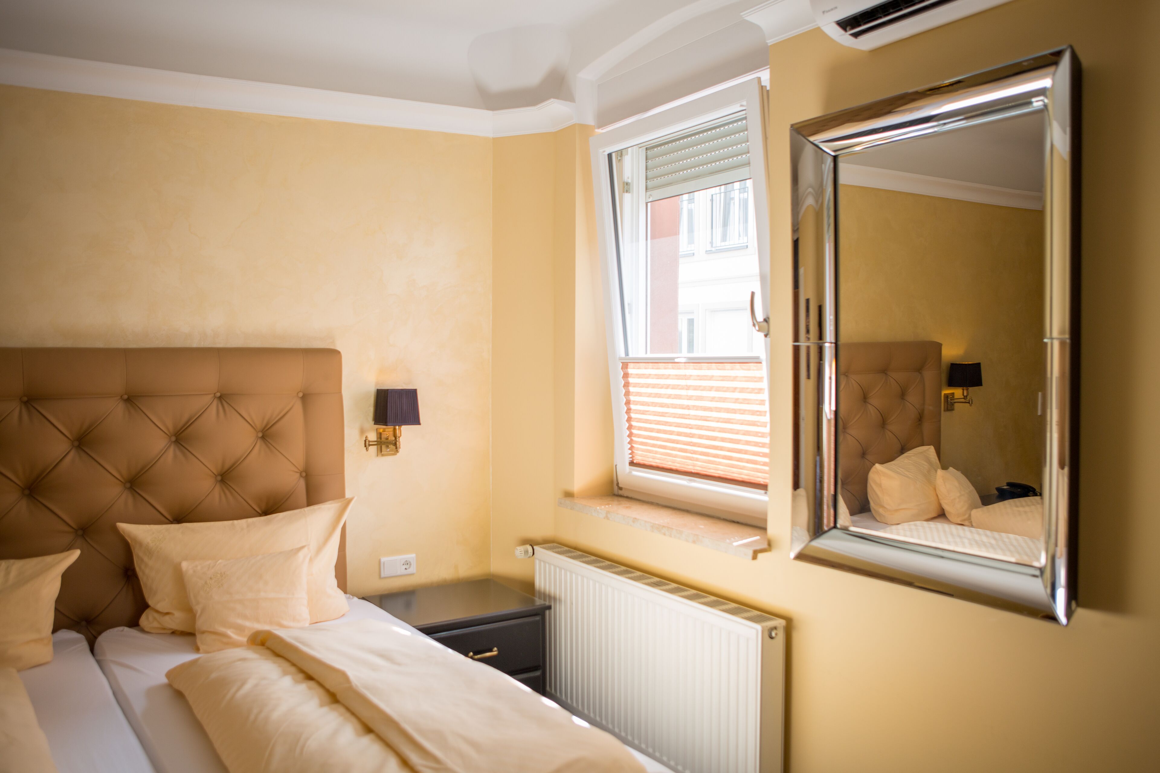 A bedroom with a tufted headboard, a large mirror, a window with blinds, and a bedside table with a lamp.