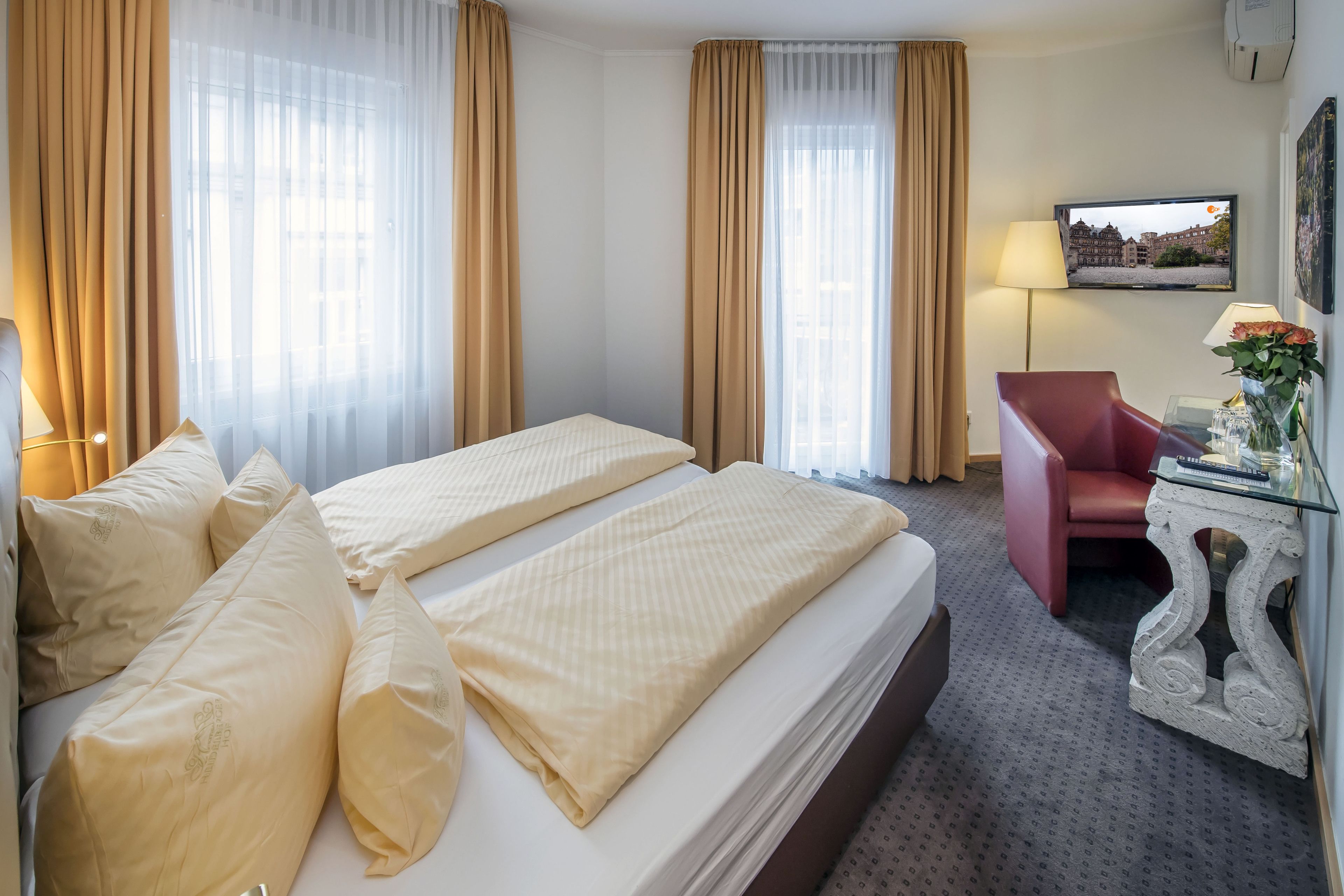A hotel room with a double bed, two bedside tables, a red armchair, a TV on the wall, and a window with curtains.