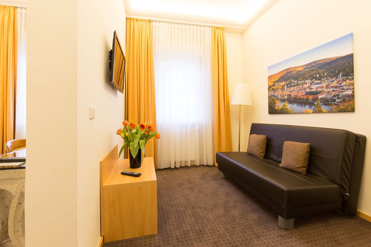 A hotel room with a sofa, a small table with a vase of flowers, a painting on the wall, and a window with curtains.
