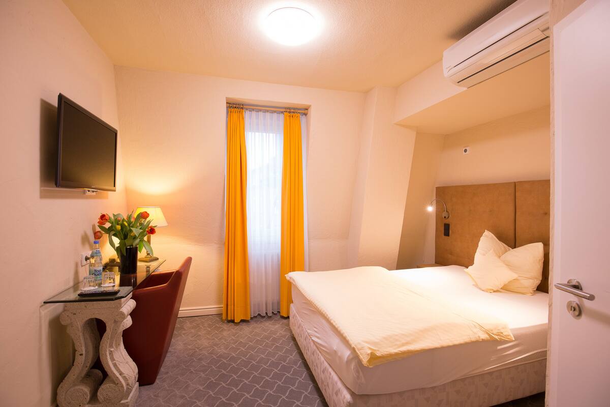 A hotel room with a bed, a TV mounted on the wall, a desk with a chair, and a window with curtains.