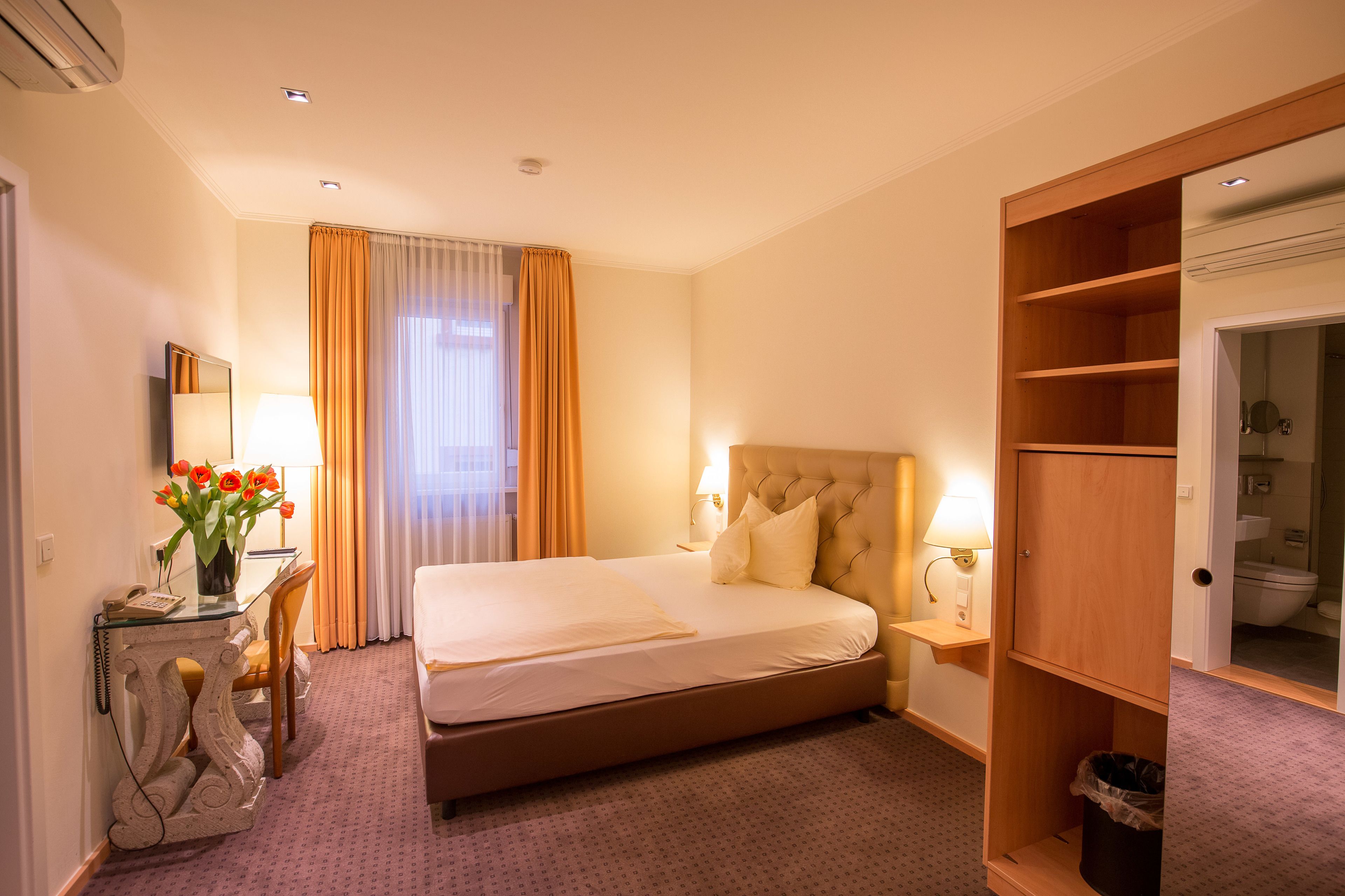 Quarto de hotel com cama, mesa de cabeceira, abajur e guarda-roupa.