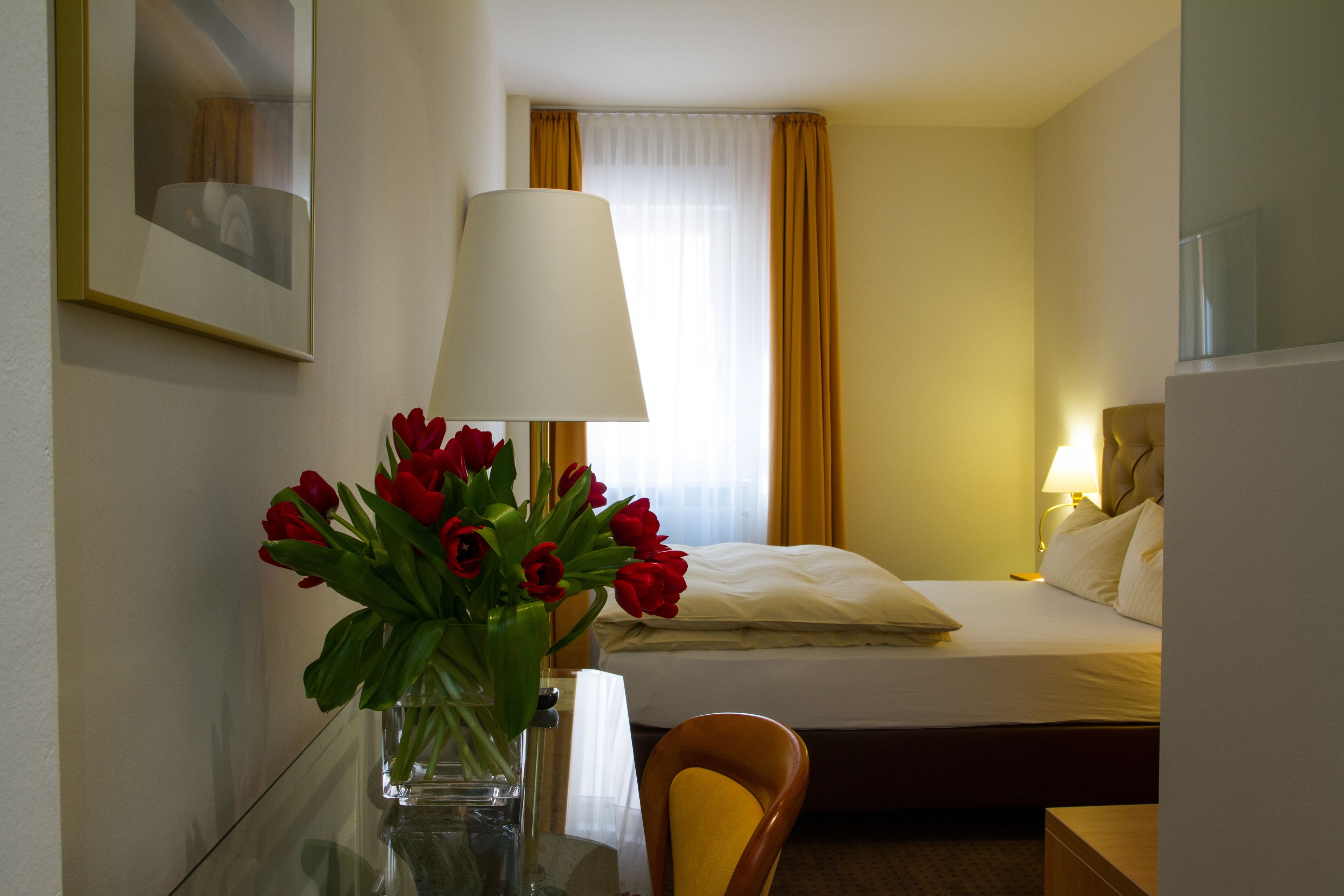 Una habitación de hotel con una cama, una mesita de noche con lámpara, una silla y un florero con flores rojas.