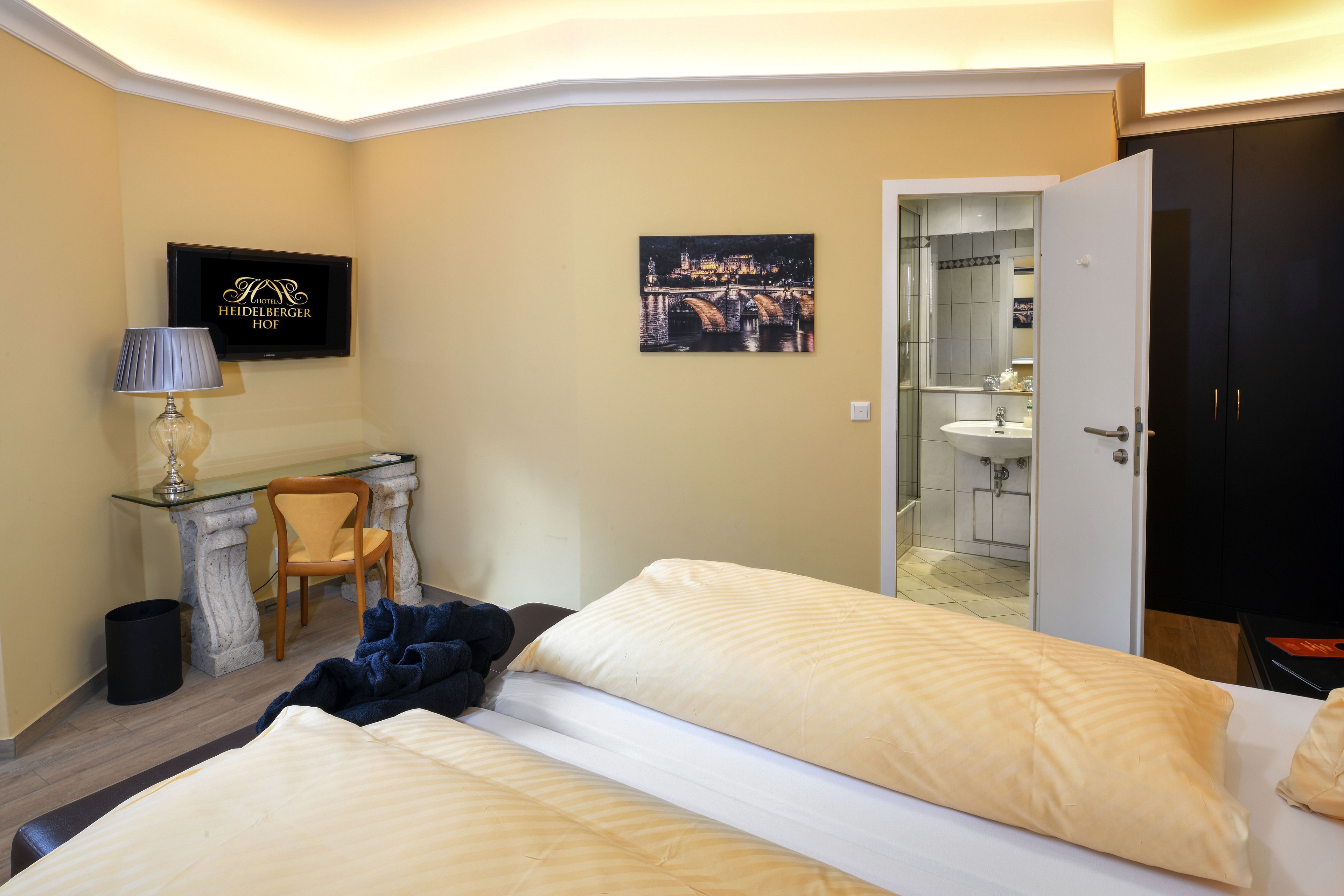 A hotel room with a bed, a desk, a chair, a TV, and a bathroom visible through an open door.