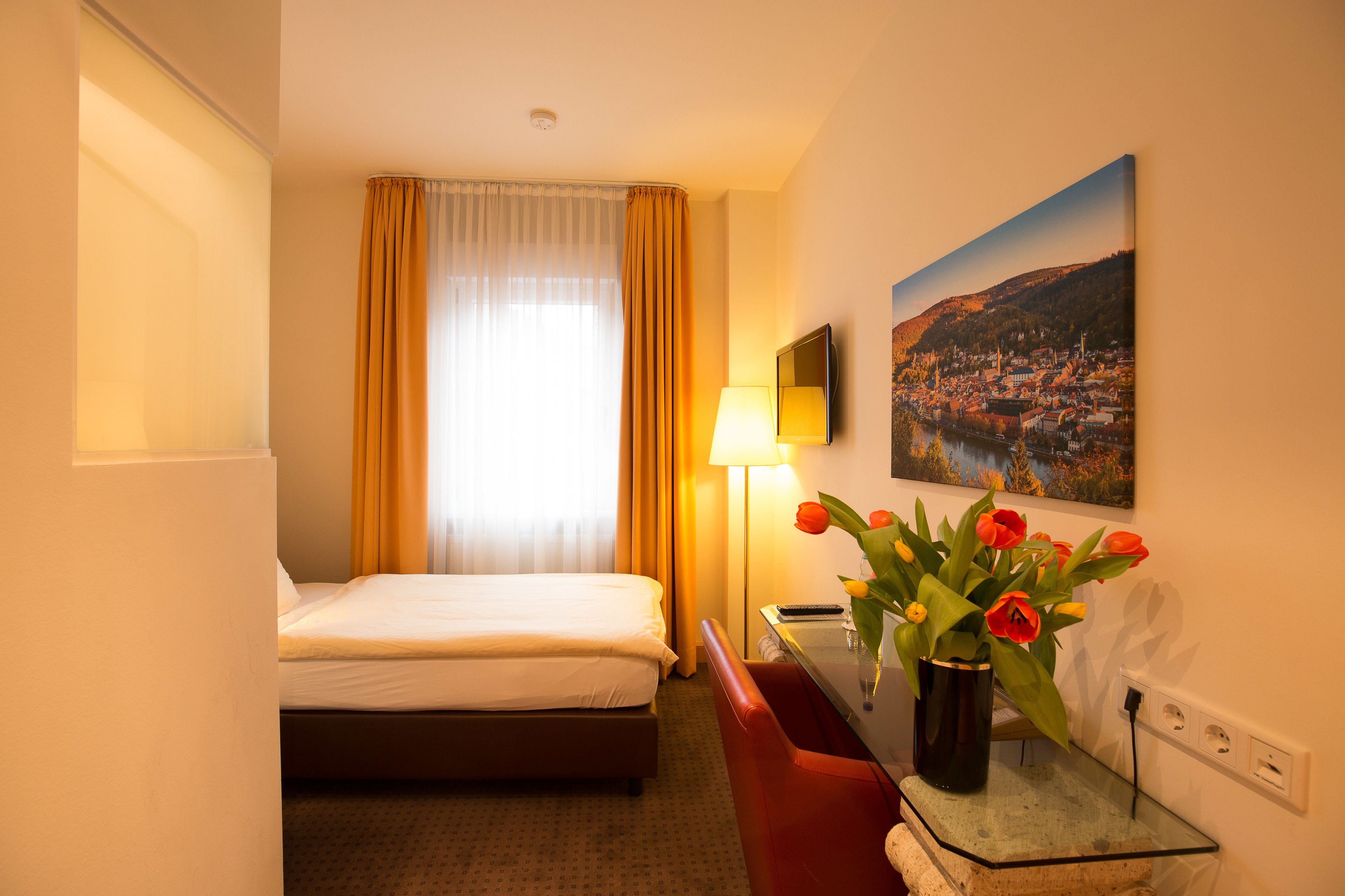 Habitación de hotel con cama, escritorio con lámpara, jarrón con flores y un cuadro en la pared.