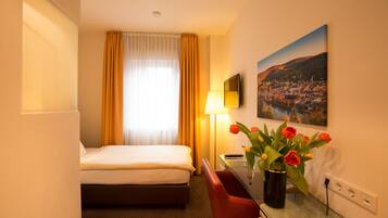 Habitación de hotel con cama, escritorio con lámpara, jarrón con flores y un cuadro en la pared.