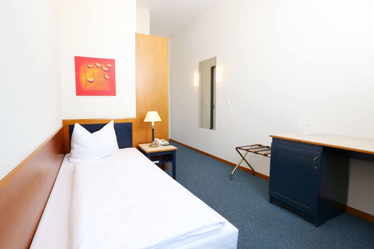 A hotel room with a bed, a desk, a lamp, a phone, a mirror, and a small cabinet.