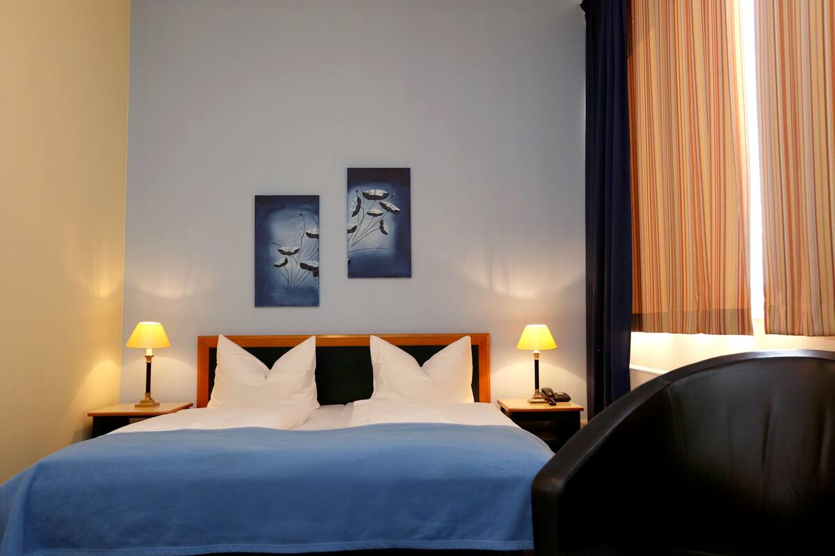 A hotel room with a bed, two bedside lamps, a nightstand, a chair, and a window with curtains.