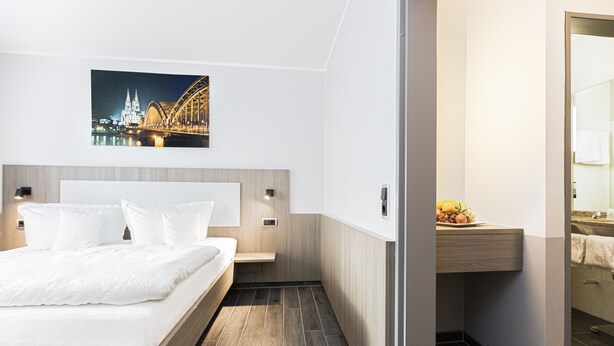 A modern hotel room with a large bed, a wooden headboard, a night view of a bridge, and a small bathroom area. - Hotel Haus Fück (Leverkusen)