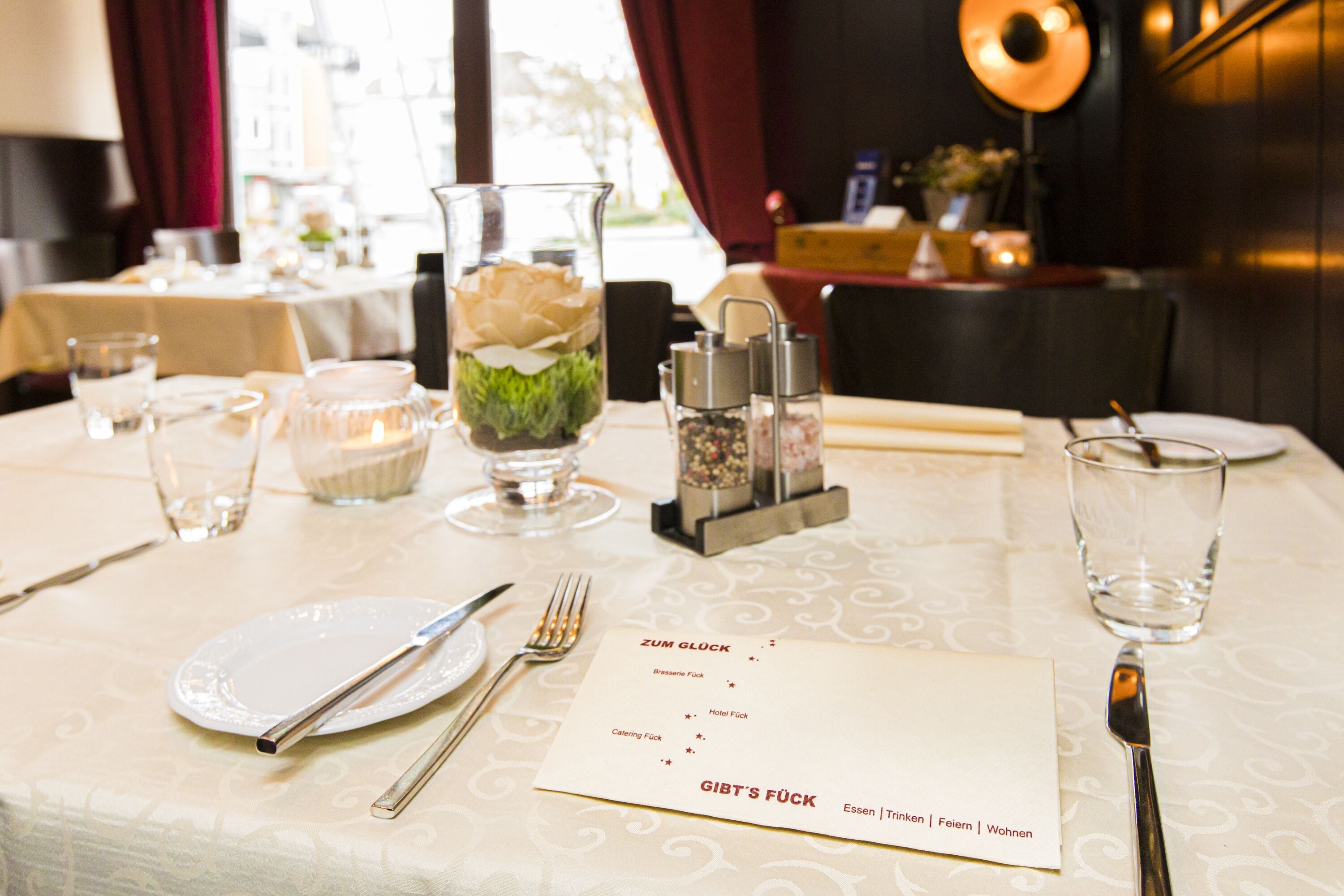 A dining table set with a menu, glasses, cutlery, and a decorative vase with a flower.