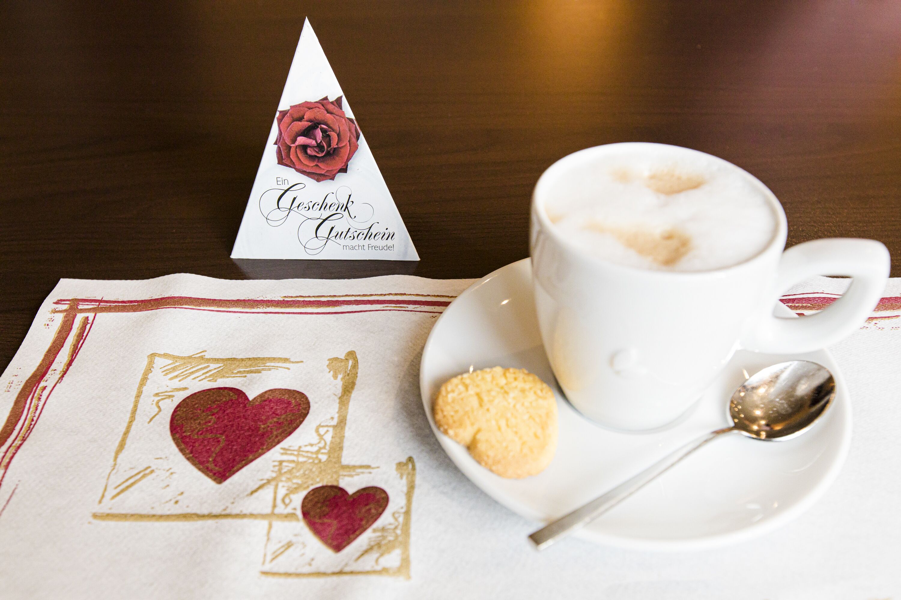 A cup of coffee with a heart-shaped foam on top, a biscuit, and a napkin with a heart design.