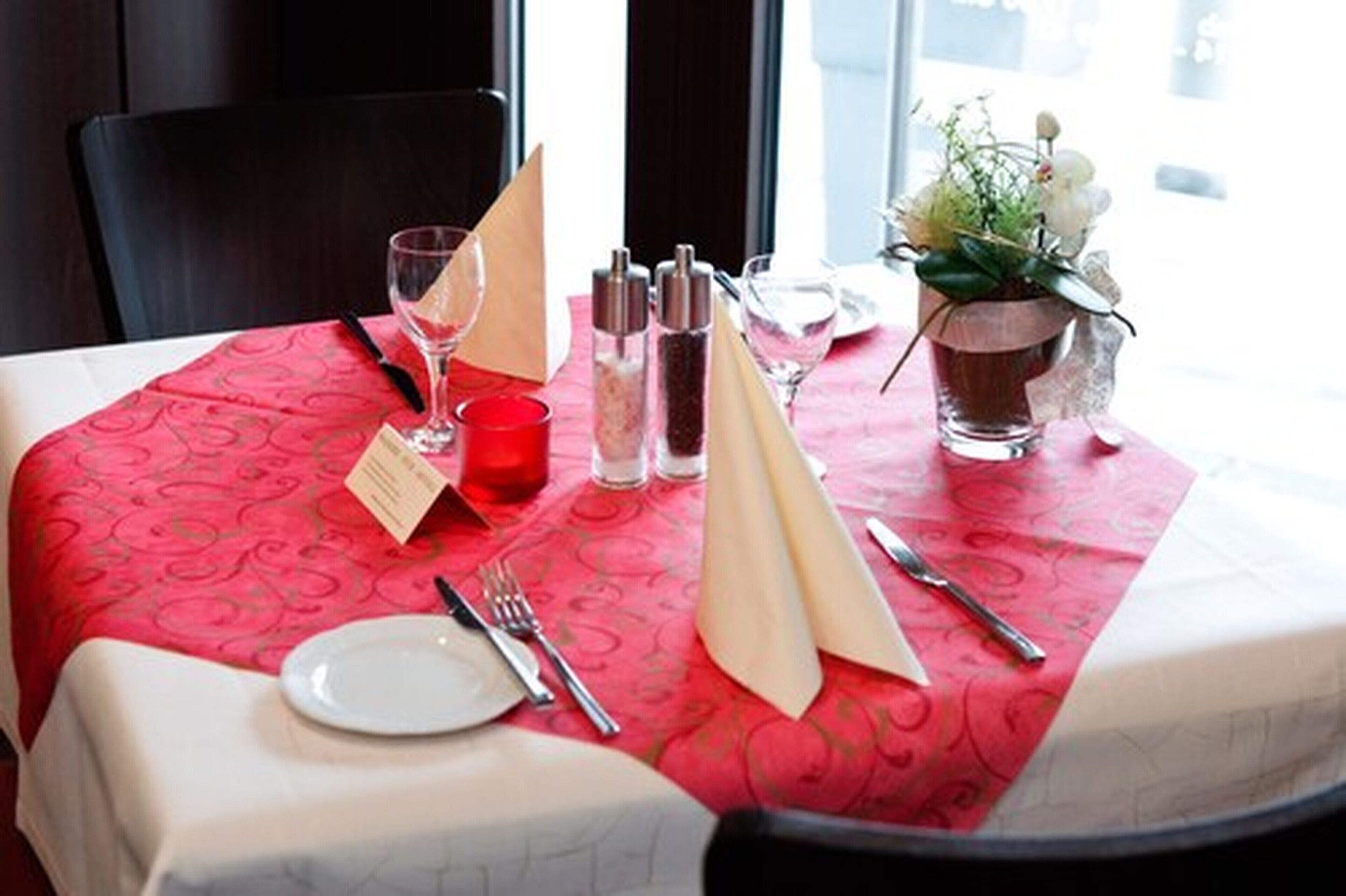 A table set for dining with a red tablecloth, a menu, salt and pepper shakers, a vase with flowers, and a candle.