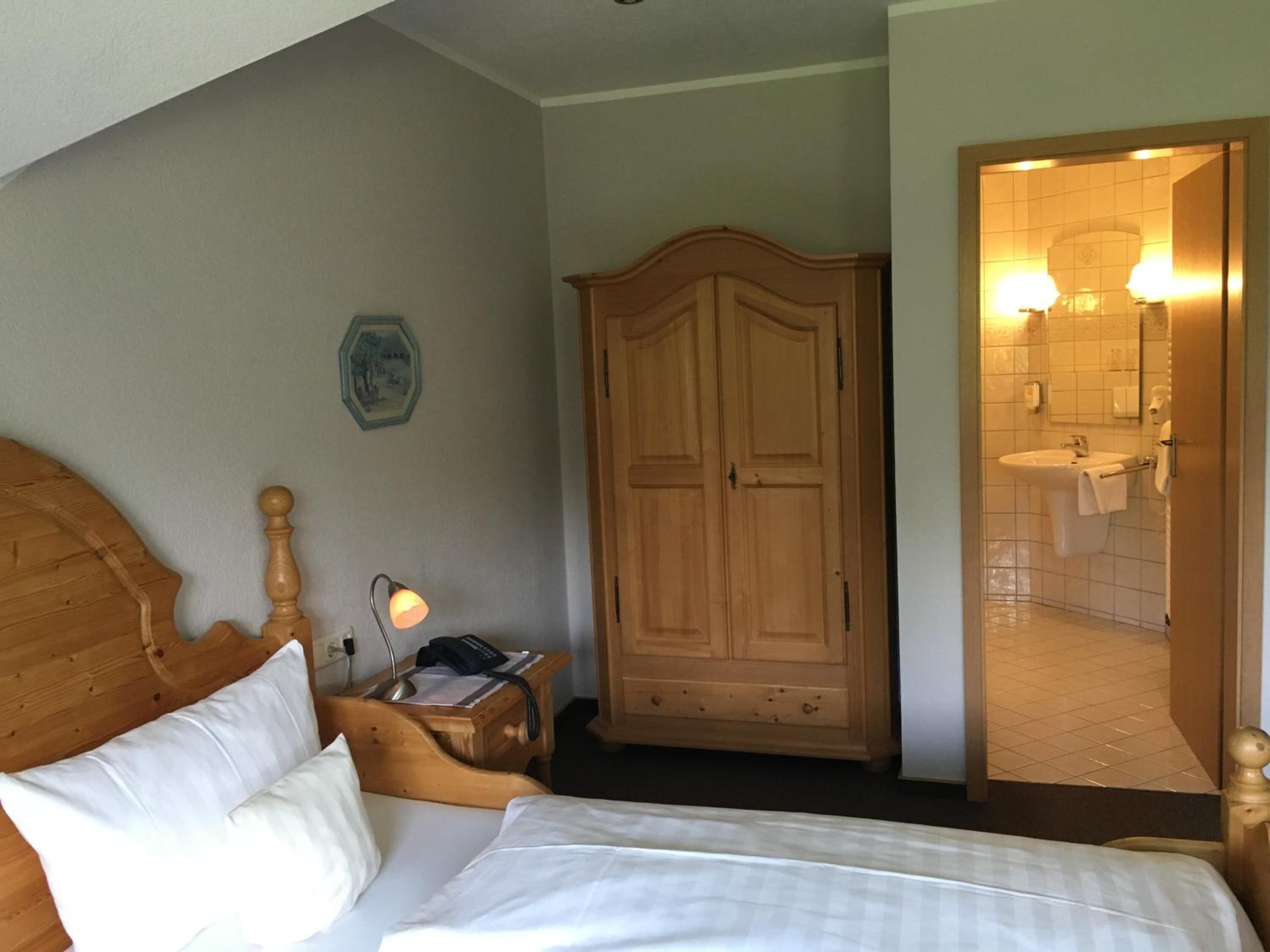 A bedroom with a wooden bed, a nightstand with a lamp, a wooden wardrobe, and an open door to a bathroom with a sink and mirror.