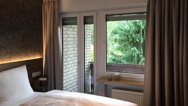A bedroom with a large bed, a window with curtains, a brick wall, and a view of greenery outside. - Landhaus Pension Göke (Hoevelhof)