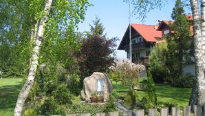 A stone monument with a plaque in a landscaped garden, a wooden fence, and a building with a red roof in the background. - Landhaus Pension Göke (Hoevelhof)