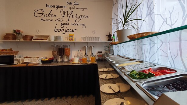 Montaje de bufé con diferentes alimentos y bebidas, una planta en maceta y una pared con saludos matutinos en varios idiomas.