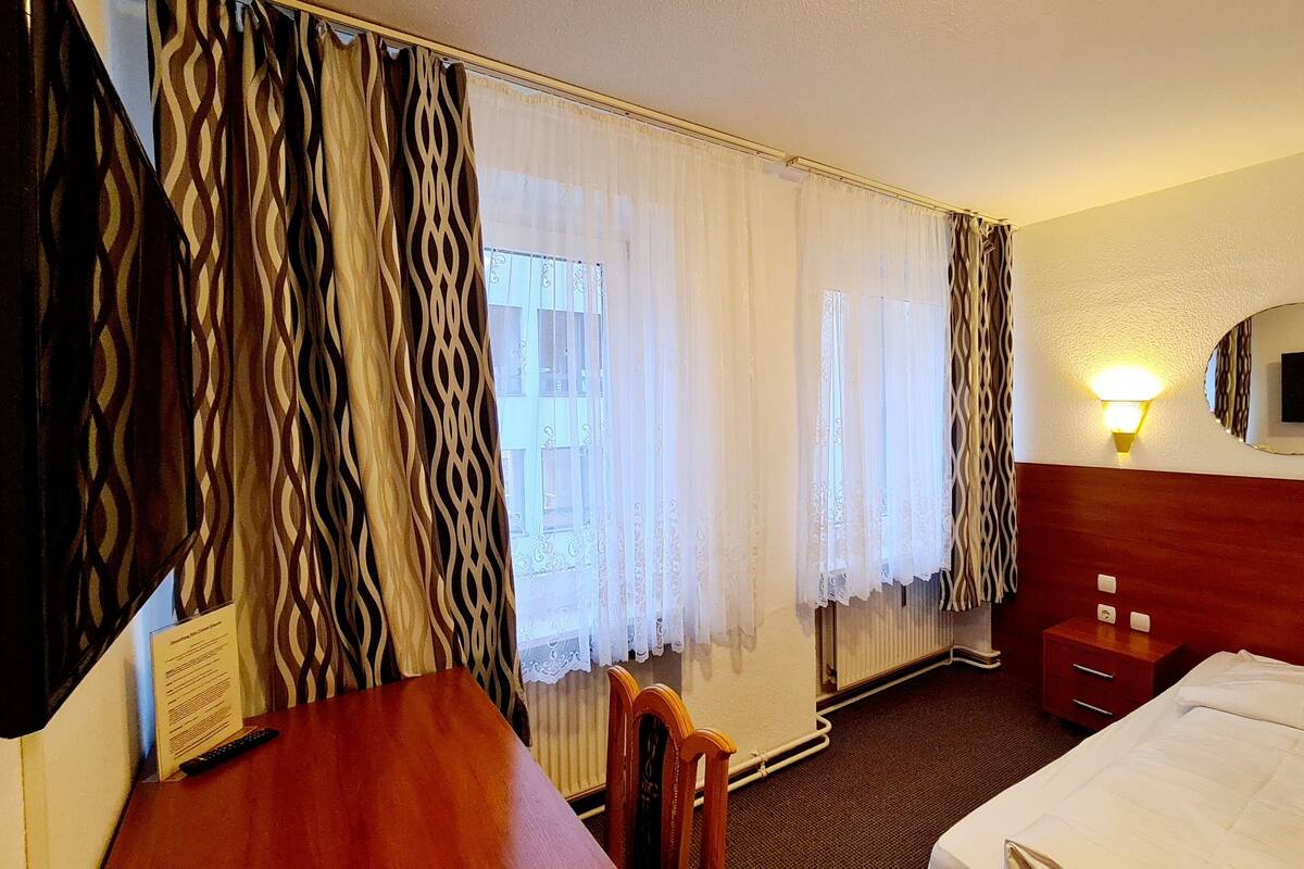 Hotelkamer met een bed, bureau, stoel, nachtkastje, spiegel en raam met gordijnen.