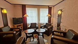 Habitación de hotel con televisor, dos sillas, una mesita y una ventana con cortinas.
