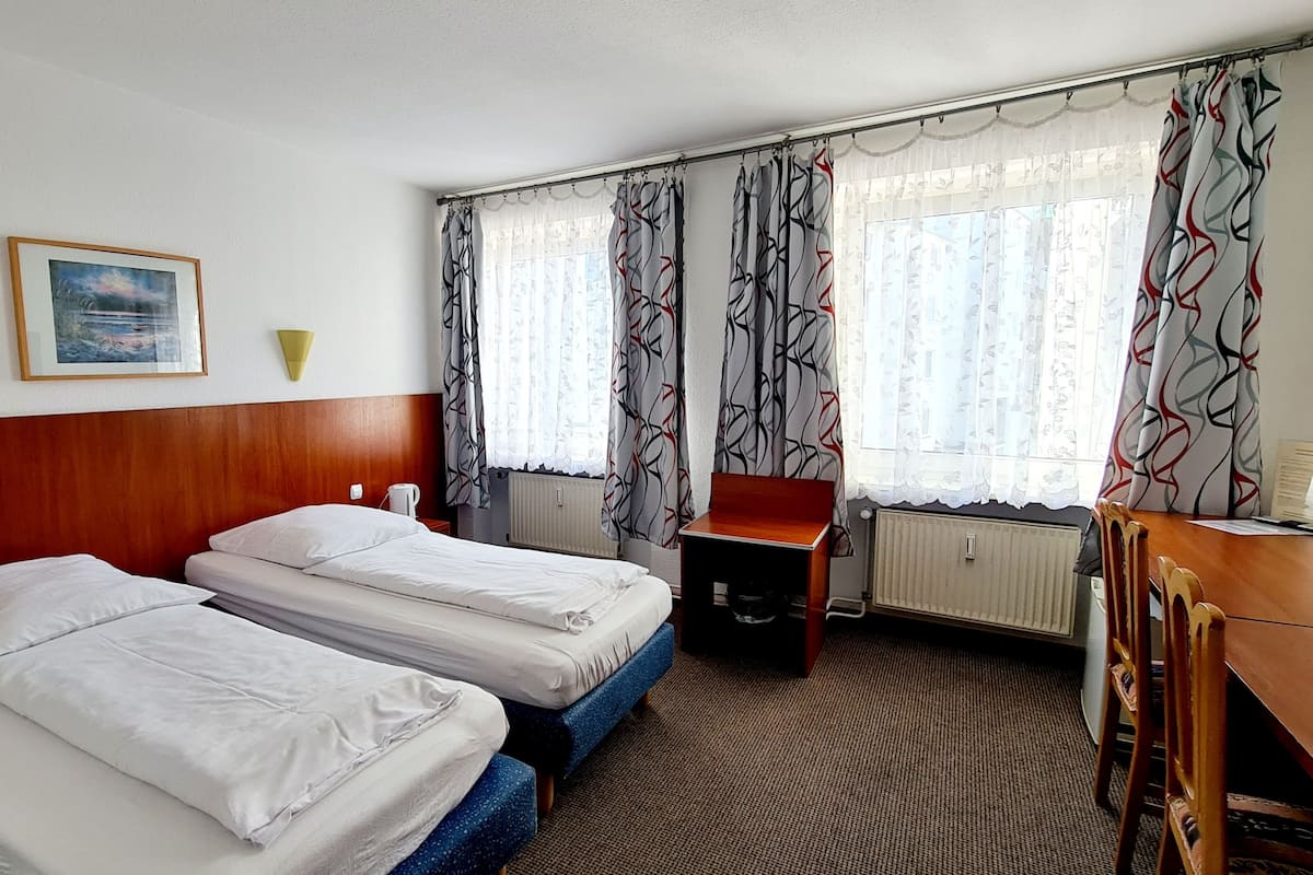Een hotelkamer met twee enkele bedden, een houten hoofdbord, een klein bureau, een stoel, een radiator en een raam met decoratieve gordijnen.