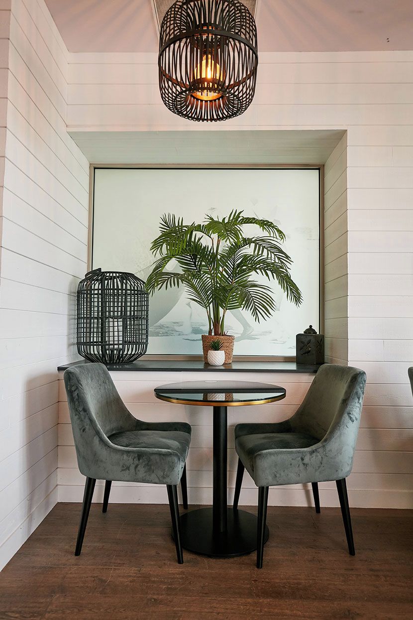 Une pièce avec une table ronde, deux chaises, une plante en pot, un luminaire suspendu et une fenêtre donnant sur un paysage.