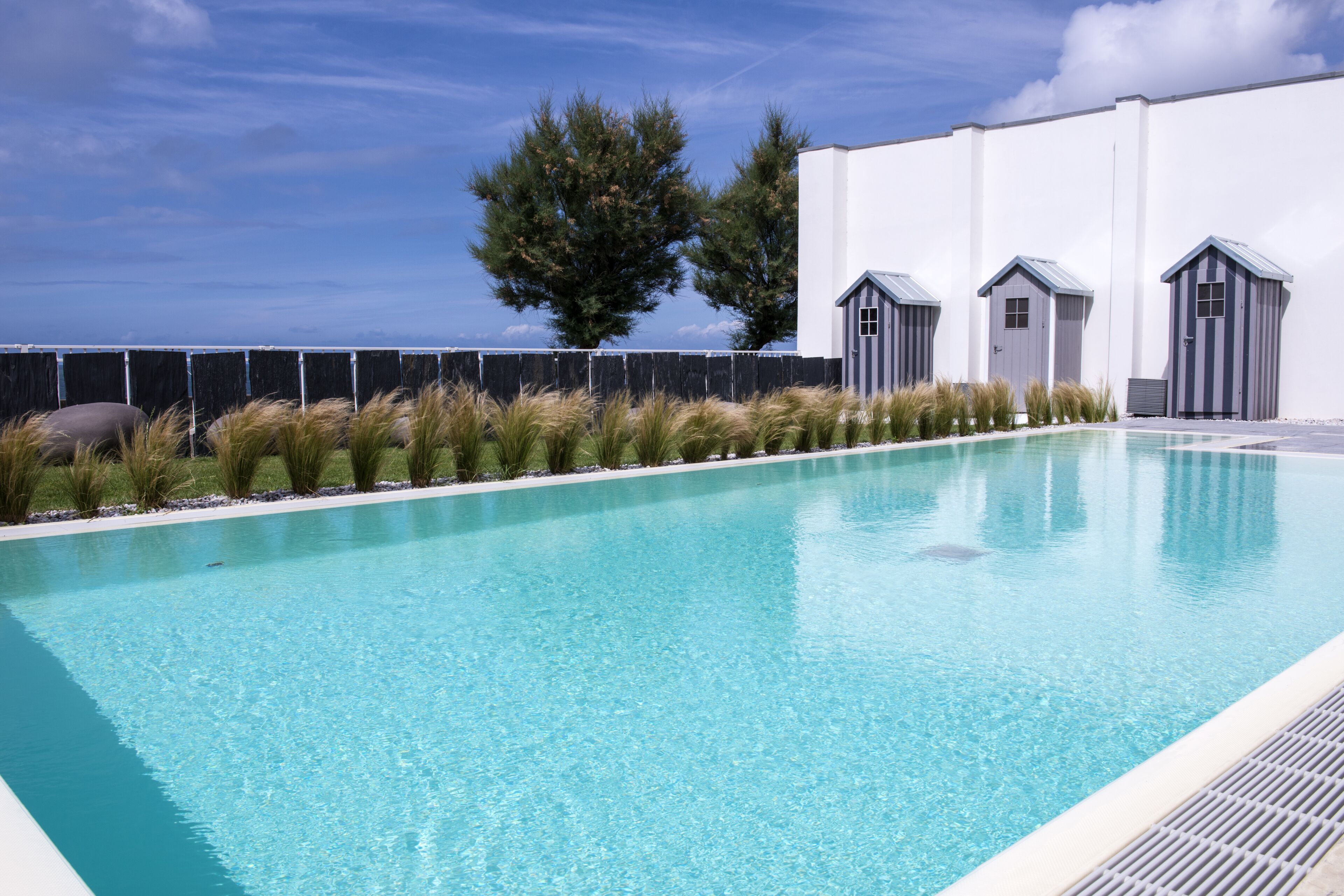 Une piscine rectangulaire à l’eau d’un bleu cristallin, entourée d’un petit muret en pierre et bordée d’une rangée de petits abris.