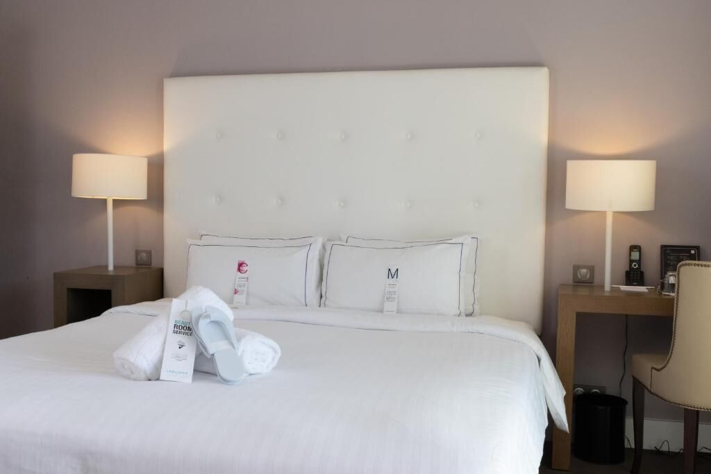 A neatly made bed with pillows labeled 'C' and 'M', a bedside table with a lamp, and a chair.