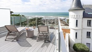 Un balcón con dos sillones de descanso y una mesa, con vistas a una playa y un edificio blanco con una torre.