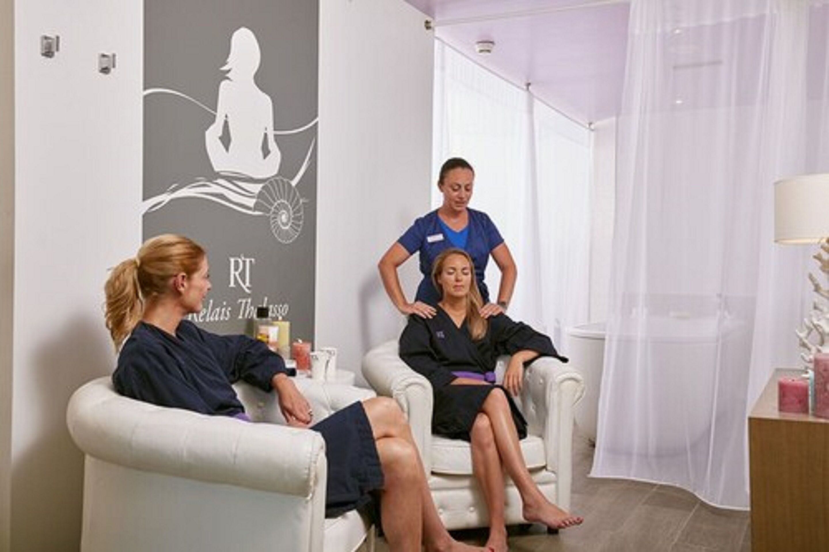 Un salón de spa con dos clientes sentados en sillones, un terapeuta detrás de uno de ellos, y una pared con la silueta de una mujer meditando.