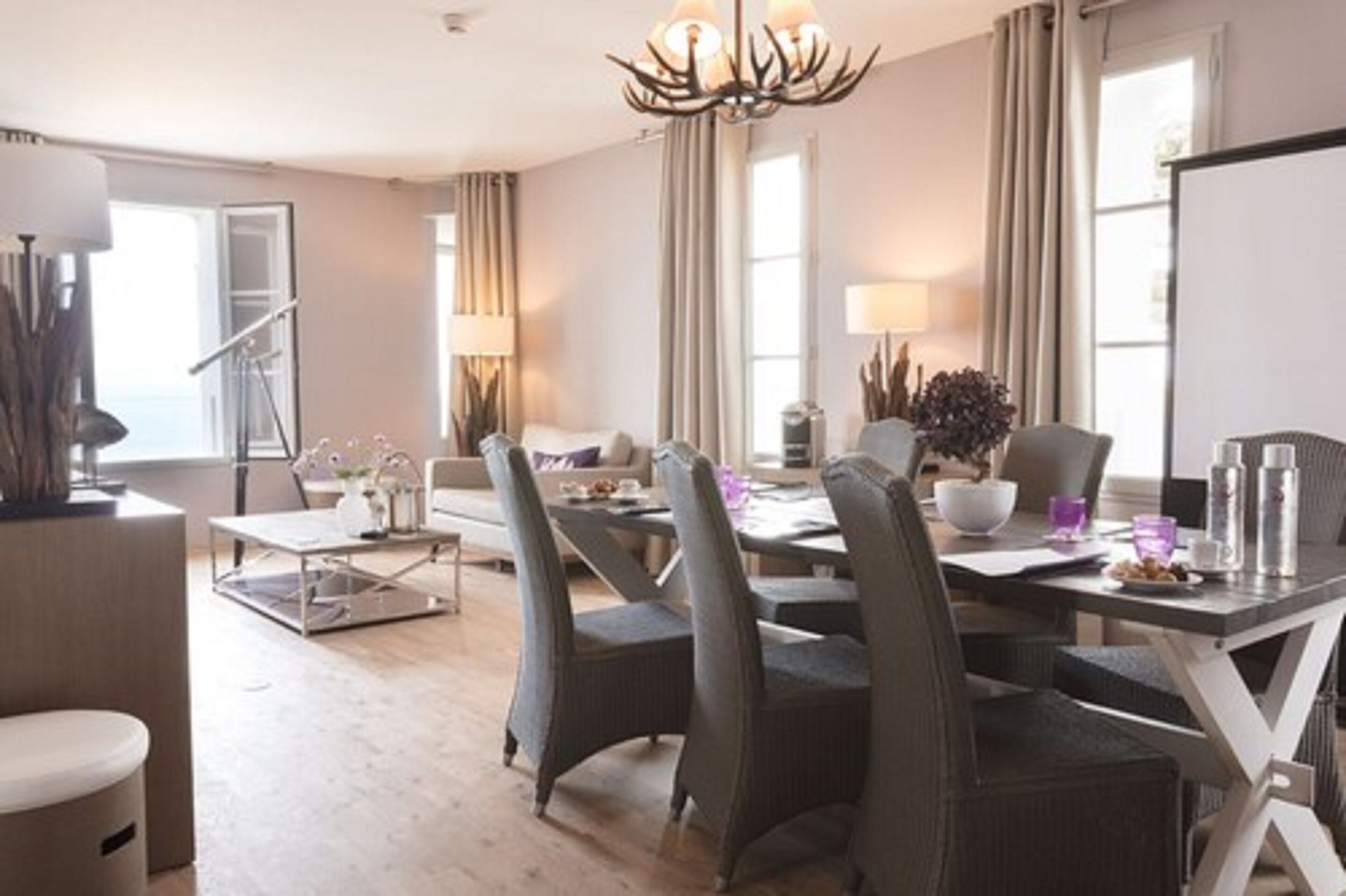 Uma sala de jantar moderna com uma mesa grande, cadeiras e um candelabro. Há um sofá e uma mesinha de centro com objetos decorativos.